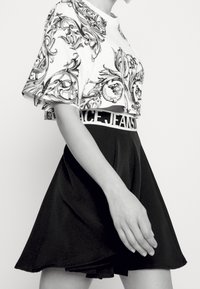 Zkrácený bílý top s černým květinovým vzorem a rozšířenou černou sukni s bílým pasem označeným "PEACE JEANS." Hladké, lehké materiály.