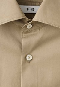 Camisa beige con cuello clásico, con dos botones blancos y una textura de tela suave. La etiqueta dice "MNG."