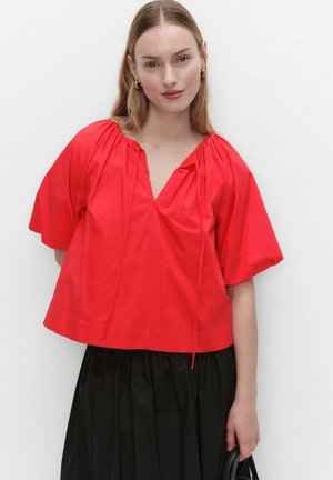 Nainen, jolla on löysä punainen pusero, jossa on lyhyet liehupuffihihat, ja musta hame, pitäen mustaa käsilaukkua yksivärisen valkoisen taustan edessä.