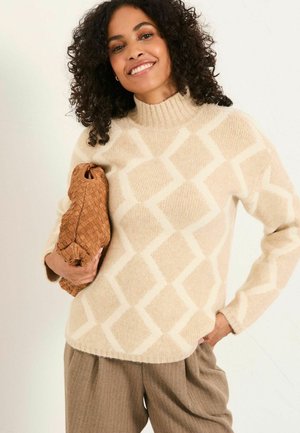 Lächelnde Frau mit lockigem Haar, die einen beigen Pullover mit Diamantmuster und braune Hosen trägt und eine geflochtene braune Clutch-Tasche hält.