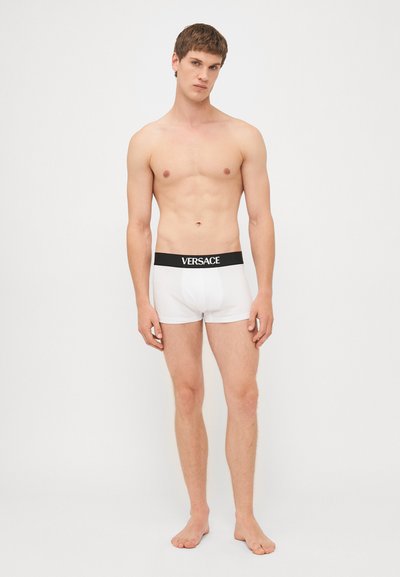 Weiße Baumwollboxershorts mit einem schwarzen Gummibund, der das Versace-Logo in Weiß trägt. Glatte Textur, enganliegendes Design, mit Länge bis zur Mitte des Oberschenkels.