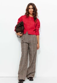 Cardigan rouge à côtes avec des détails de boutons, associé à un pantalon large à carreaux marron. Sac marron foncé, chaussures noires brillantes. Fond blanc uni.