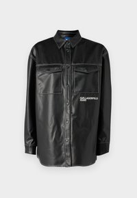 Giacca camicia in ecopelle nera con chiusura a bottoni, due tasche sul petto, cuciture bianche a contrasto e un logo sul lato sinistro.