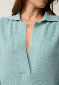 Vaaleansininen neulepaita, jossa on syvä V-pääntie ja raidoitettu kaulus. Kangas näyttää pehmeältä, sileäpintaiselta ja hienovaraisesti kiiltävältä.