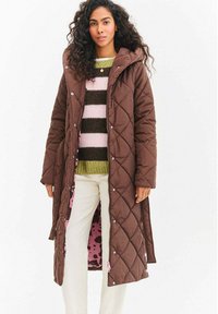 Žena má na sebe dlhý hnedý prešívaný kabát s ružovým vzorovaným podšívkou, pod ktorým má zeleno-ružový pruhovaný sveter a svetlé nohavice.