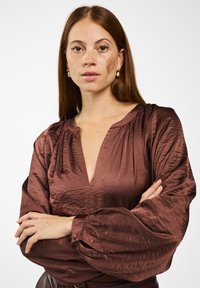 Bruine, gestructureerde blouse met een diepe V-hals en volumineuze lange mouwen. Gemaakt van glanzende stof, met geplooide accenten op de schouders.
