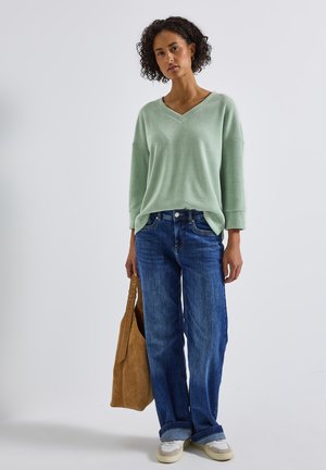 Pull en maille vert clair à col en V oversize, jeans bleu à jambes larges, sac fourre-tout en suède beige et baskets blanches. Design simple et texturé.