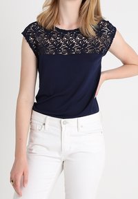 Mörkblå topp med spetsöverlägg vid axlarna, kompletterad med korta ärmar. Bärs med vita jeans som visar en avslappnad passform och en ledig stil.