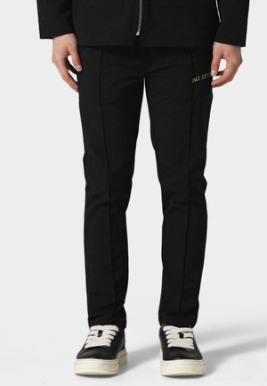 Black tapered pants with side seam detailing, featuring a 'PAS DE' gold text accent. Fabric has a smooth texture; paired with black sneakers.