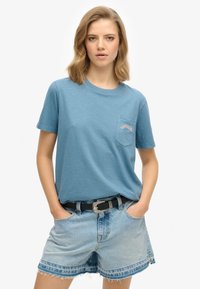 Lichtblauwe T-shirt met korte mouwen en een zak waarop "SUPERDRY" in oranje staat, gecombineerd met versleten denim shorts en een zwarte riem.