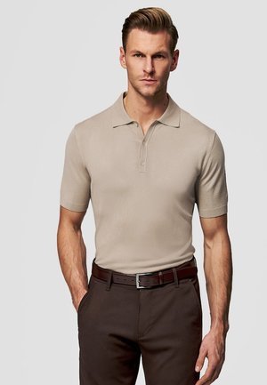 Mężczyzna o blond włosach, ubrany w beżową koszulkę polo z krótkim rękawem, ciemnobrązowe spodnie oraz dopasowany skórzany pasek, stojący na tle szarego tła.