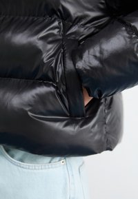 Sort puffejakke med en glat, blank overflade og en sidelomme. Ærmet er let sammensamlet, hvilket viser et let design.