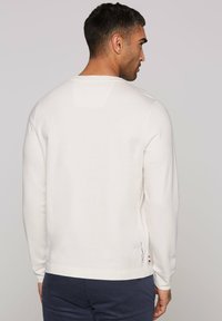 White long-sleeve shirt with a round neckline and ribbed cuffs, featuring a small brand logo at the hem and a slightly textured fabric.