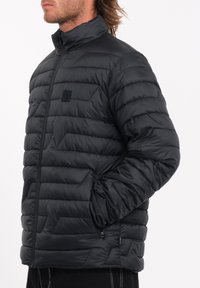 Zwarte puffy jas met een hoge kraag, voorzien van een ritssluiting en een geometrisch quiltpatroon. Textuurstof met een klein logo-embleem op de borst.