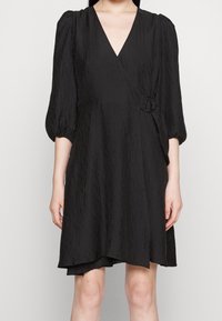 Robe noire enveloppante en tissu texturé, dotée d'un décolleté en V, de manches bouffantes et d'un détail en D-ring sur le côté pour l'ajustement.