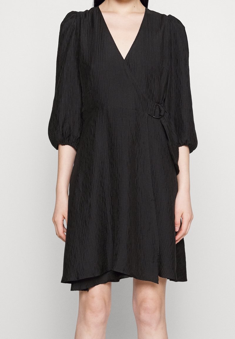 Robe noire enveloppante en tissu texturé, dotée d'un décolleté en V, de manches bouffantes et d'un détail en D-ring sur le côté pour l'ajustement.