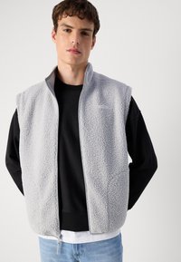 Lys grå fleece vest med frontlås, høy krage og ermeløs design. Bæret over en svart langermet skjorte. Teksturert overflate.