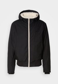 Giacca nera isolata con cappuccio, foderata in pelliccia sintetica bianca e con zip frontale. Presenta polsini e orlo a coste, con dettaglio del logo sul petto.