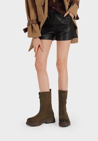 Stivaletti in suede marrone alla caviglia con zip laterale, sole spesso e texture opaca, abbinati a pantaloni corti in pelle nera e una giacca beige.