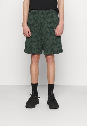 Personne portant un short Nike camouflage vert foncé, des chaussettes noires et des chaussures de sport noires, debout devant un fond clair uni.