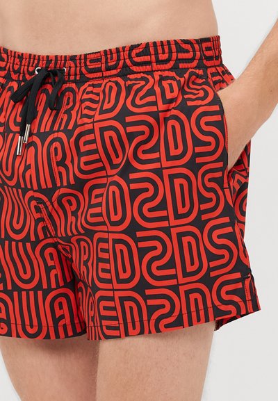 Homme portant un short de bain noir avec un motif de logo rouge audacieux DSQUARED2, taille élastique, cordon de serrage, et main dans la poche droite.