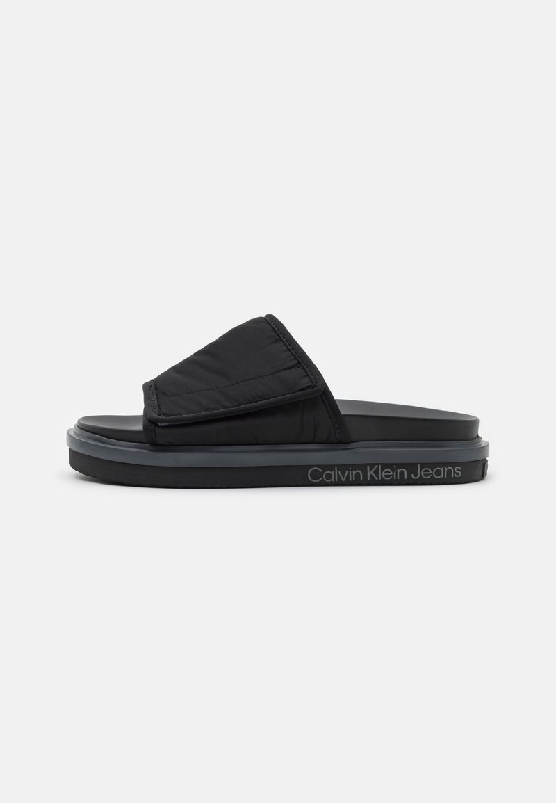 Sandale slide noire Calvin Klein Jeans avec bride supérieure rembourrée et semelle épaisse, vue de côté sur fond blanc.