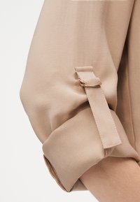 Manche beige à manches longues retroussées avec sangle en tissu et fermeture à boucle dorée au-dessus de l'avant-bras sur un bras à peau claire.