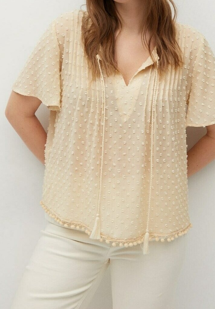 Woman wearing a beige textured short-sleeve blouse with tassel ties and cream pants, standing with hands behind her back.