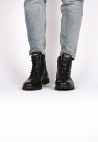 Botas de cuero negras con cordones en la parte delantera y cremallera lateral, con una suela gruesa y acabado texturizado. Llevadas con jeans de color azul claro.