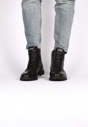 Stivali neri in pelle con allacciatura frontale e zip laterale, caratterizzati da una suola spessa e una finitura testurizzata. Indossati con jeans azzurri chiari.