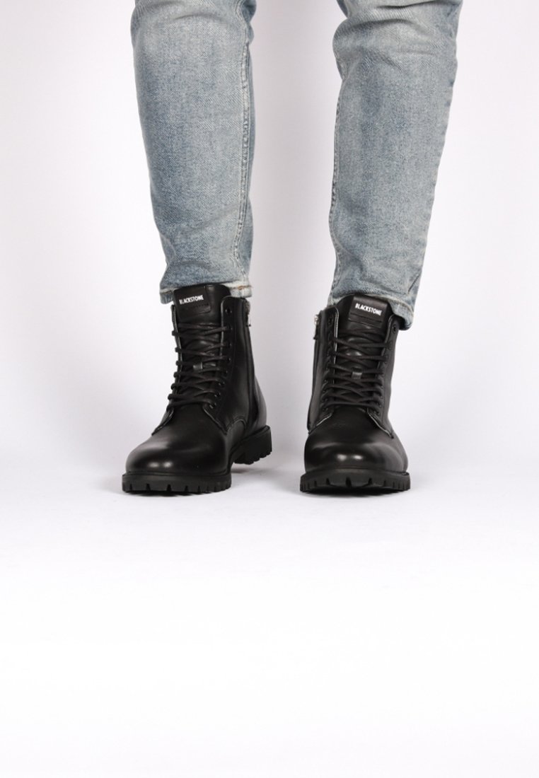 Botas de cuero negras con cordones en la parte delantera y cremallera lateral, con una suela gruesa y acabado texturizado. Llevadas con jeans de color azul claro.