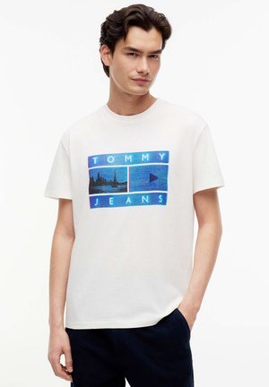 Młody mężczyzna w białym T-shircie Tommy Jeans z niebieskim nadrukiem i ciemnych spodniach, stoi z jedną ręką w kieszeni na białym tle.