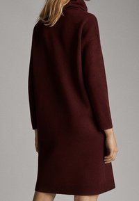 Bordeaux värinen ylipitkä neulemekko, jossa on korkea kaulus, pitkät hihat ja suora siluetti. Kangas vaikuttaa pehmeältä ja teksturoidulta.