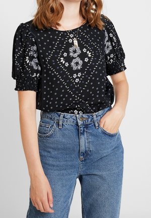 Vrouw draagt een blauwe high-waist jeans en een zwart bloesje met pofmouwen en witte bloemenpatronen, een zilveren ketting, met één hand in de zak.