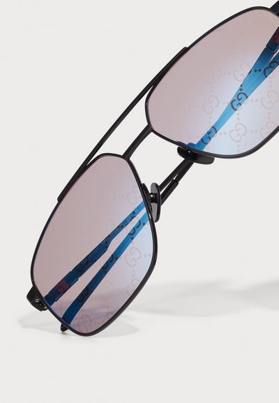 Czarne okulary przeciwsłoneczne z metalową oprawą i delikatnie przyciemnianymi soczewkami, odbijającymi niebieski pasek z powtarzającymi się logo Gucci.