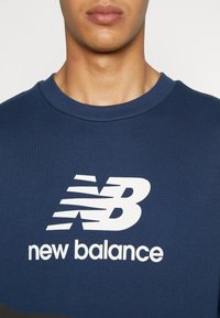 Homme portant un t-shirt col rond bleu marine avec le logo New Balance blanc et le texte centrés sur la poitrine.