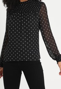 Blouse noire à manches longues avec encolure smockée, tissu transparent et motif en diamants argentés. Les poignets comportent des détails élastiques pour un ajustement optimal.