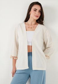 Cardigan aperto beige in lino con maniche a tre quarti e dettagli a frange ai polsini, abbinato a una maglietta bianca e pantaloni blu.