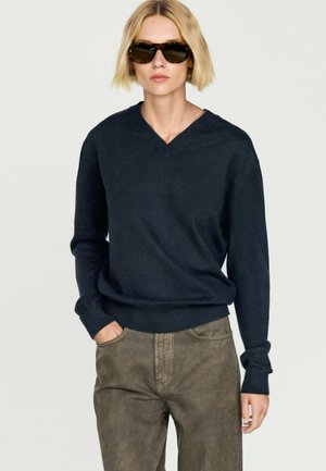 Person mit kurzen blonden Haaren, die eine Sonnenbrille, einen marineblauen V-Ausschnitt-Pullover und eine braune Hose trägt, steht mit einer Hand in der Tasche vor einem schlichten Hintergrund.