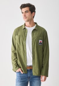 Olive green corduroy shirt with vertical stripes, button closure, chest pocket, and a patch on the left side, worn over a white T-shirt.
