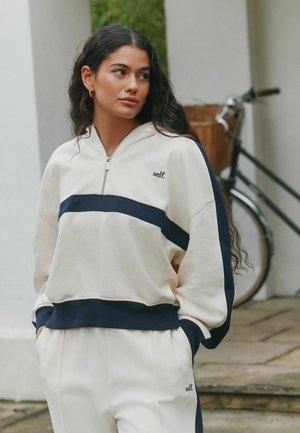 Lichtcrème hoodie met marineblauwe strepen, kwart-zip ontwerp en geborduurd "self." logo. Gecombineerd met bijpassende crème sweatpants.