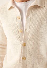 Helles cremefarbenes Strickjacke aus strukturiertem Strickstoff, mit V-Ausschnitt, langen Ärmeln und fünf gelben Knöpfen vorne.