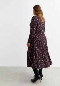 Vestido negro de manga larga con un patrón abstracto en rosa, diseño fluido y dobladillo acampanado, combinado con botas negras hasta la rodilla.