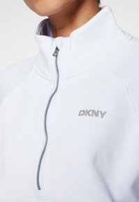 Világoskék, negyedzipzáras, magas gallérú pulóver. Ezüst cipzárral és strasszkövekkel díszített "DKNY" logóval rendelkezik a mellkasán.