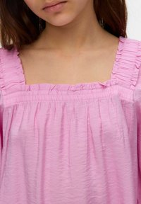 Top rosa con escote cuadrado, que presenta bordes con volantes y una tela texturizada, cayendo de manera suelta con detalles fruncidos a lo largo del cuerpo.