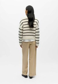 Megzta kreminė megztinis su tamsiai rudos spalvos juostelėmis, trumpintas dizainas ir gofruotas apvadas, derinamas su šviesiai rudos spalvos plačiomis kelnėmis ir juodais batais.