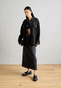 Chemise-robe noire oversize avec boutons-pressions, débardeur court, jupe maxi en denim, et mocassins noirs avec un nœud décoratif. Lisse et texturé.