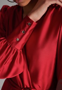 Robe en satin rouge avec des manches bouffantes. Présente des boutons argentés sur les poignets et une ceinture à la taille. Texture lisse avec un léger éclat.