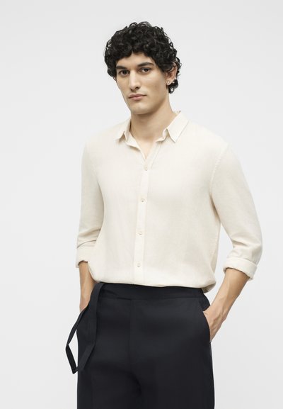 Νέος άνδρας με σγουρά μαύρα μαλλιά, που φοράει κρεμ πουκάμισο με κουμπιά και τυλιγμένα μανίκια, καθώς και μαύρο παντελόνι με τα χέρια στις τσέπες.