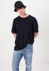 Mørkeblå kortærmet t-shirt med kontrasterende rød og hvid kant ved ærmerne, kombineret med lyseblå slidte jeans og en sort bucket hat.